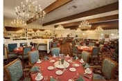 Spacious dining room with multiple round tables covered in red tablecloths, each set with plates, glasses, and napkins. The room features ornate chandeliers hanging from a ceiling with exposed wooden beams, mirrored walls, and upholstered chairs with patterned fabric.