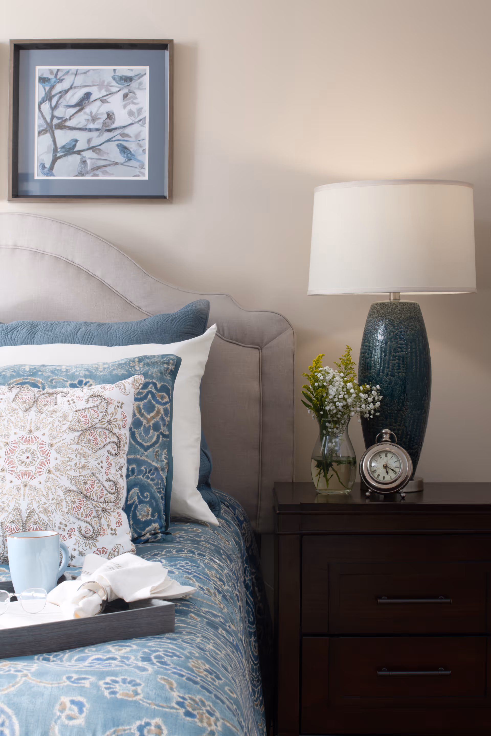 A neatly made bed with patterned blue bedding and decorative pillows next to a dark wooden nightstand holding a lamp, clock, and vase, with framed bird artwork on the wall.