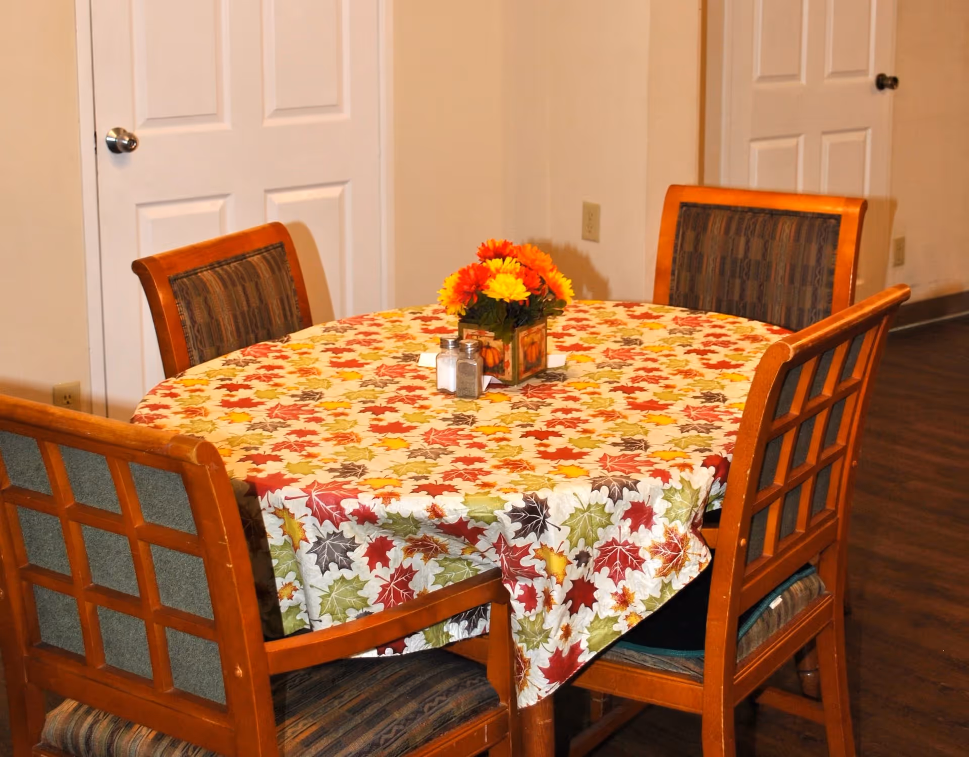 A round dining table covered with a tablecloth featuring a colorful autumn leaf pattern. The table is surrounded by four wooden chairs with cushioned seats and backs. A small floral centerpiece with orange and yellow flowers and salt and pepper shakers are placed on the table. The room has light-colored walls, two white doors with silver knobs, and a wooden floor.