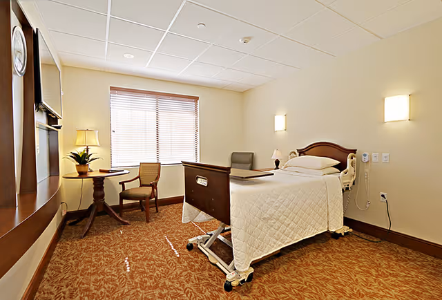 A senior living facility bedroom with a hospital-style bed covered in a white quilt, a small round wooden table with a lamp and plant, two chairs, a window with blinds, and beige walls with two wall-mounted lights.