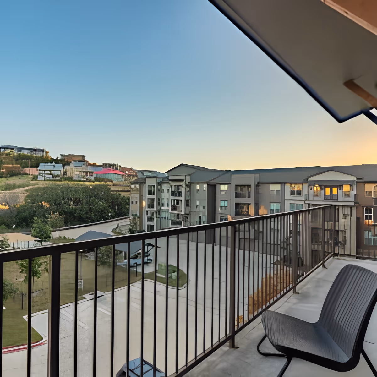 Balcony with two chairs and a metal railing overlooking apartment buildings and a hillside at sunset.