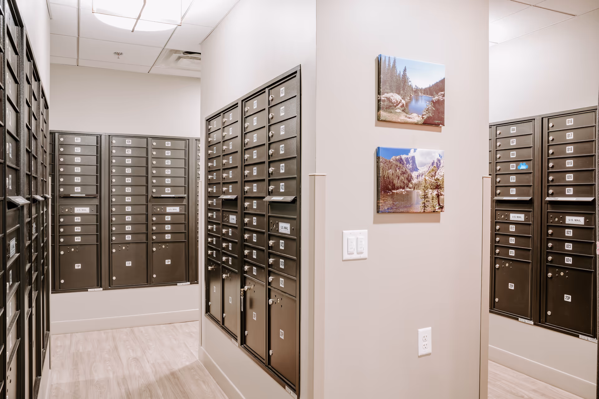 A clean interior mailroom with multiple rows of numbered black mailboxes lining the walls and two landscape photos on a central pillar.