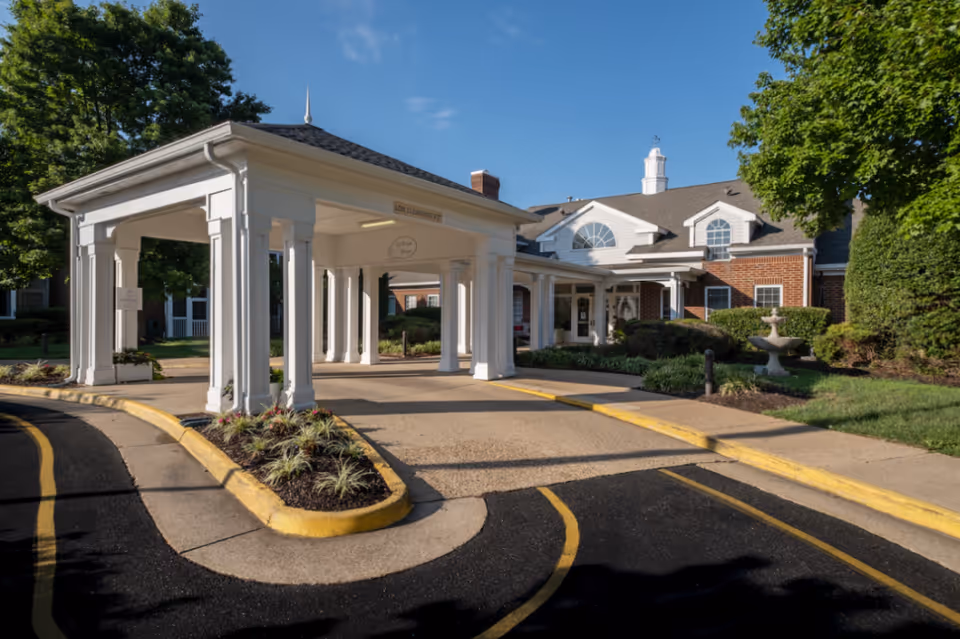 Covered porte-cochere entrance and driveway of a brick senior living building with landscaping and a fountain.