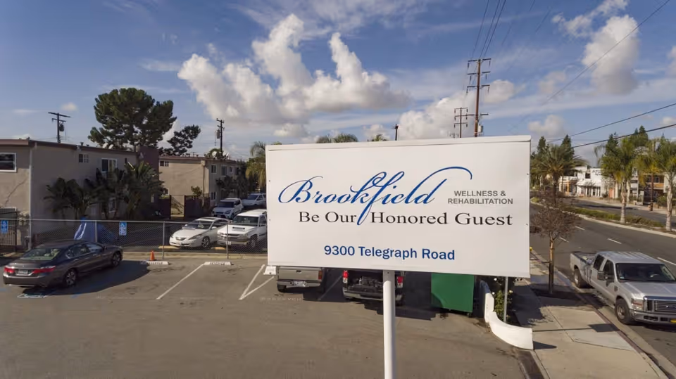 A large roadside sign reading "Brookfield Be Our Honored Guest 9300 Telegraph Road" in a parking lot with cars and buildings behind it.