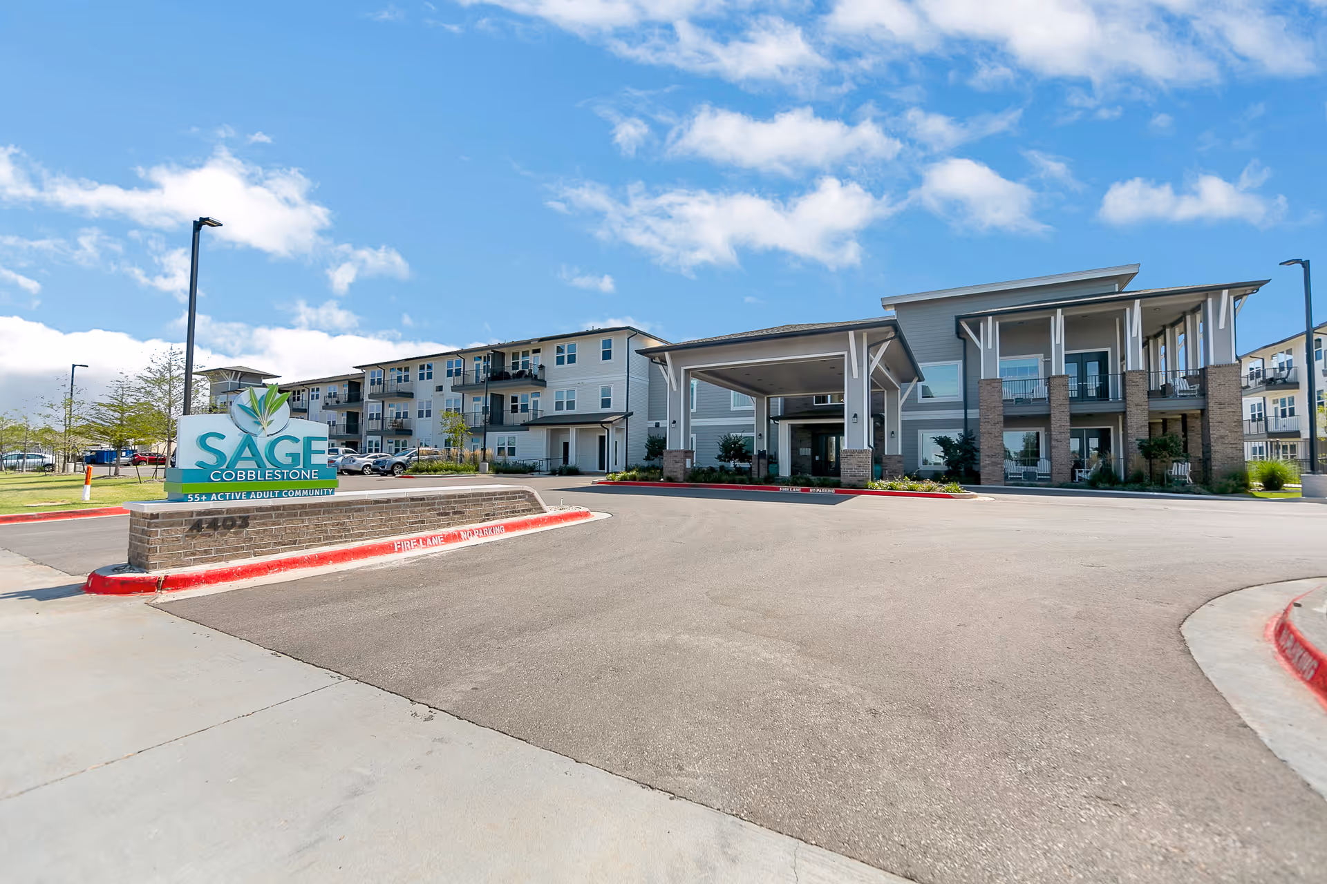 Exterior view of Sage at Cobblestone, a 55+ active adult community, showing a modern multi-story building with balconies, a covered entrance, and a sign at the front. The sky is partly cloudy and the area is well-maintained with some landscaping.