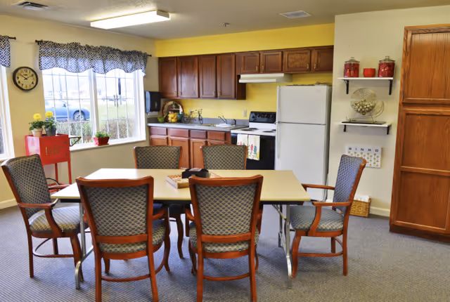 A communal dining area with a rectangular table surrounded by six upholstered chairs and a kitchenette with wooden cabinets, refrigerator and stove.