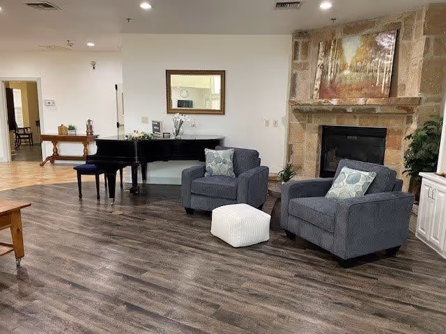 Community lounge with two gray armchairs and a white ottoman in front of a stone fireplace, a grand piano and wall art on dark wood flooring.