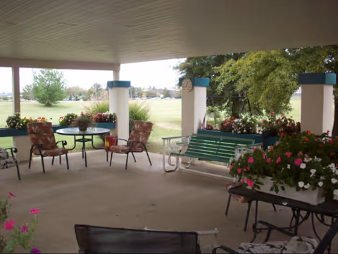Covered outdoor patio with colorful chairs, a green bench, tables, and flowering planters.