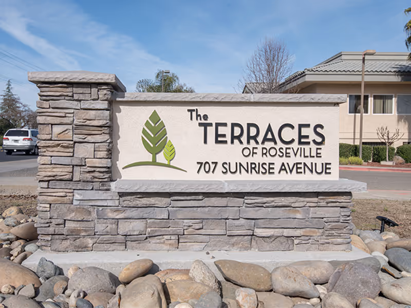 Stone sign for The Terraces of Roseville at 707 Sunrise Avenue with a logo featuring two green stylized trees, set outdoors with a building and parked car in the background.