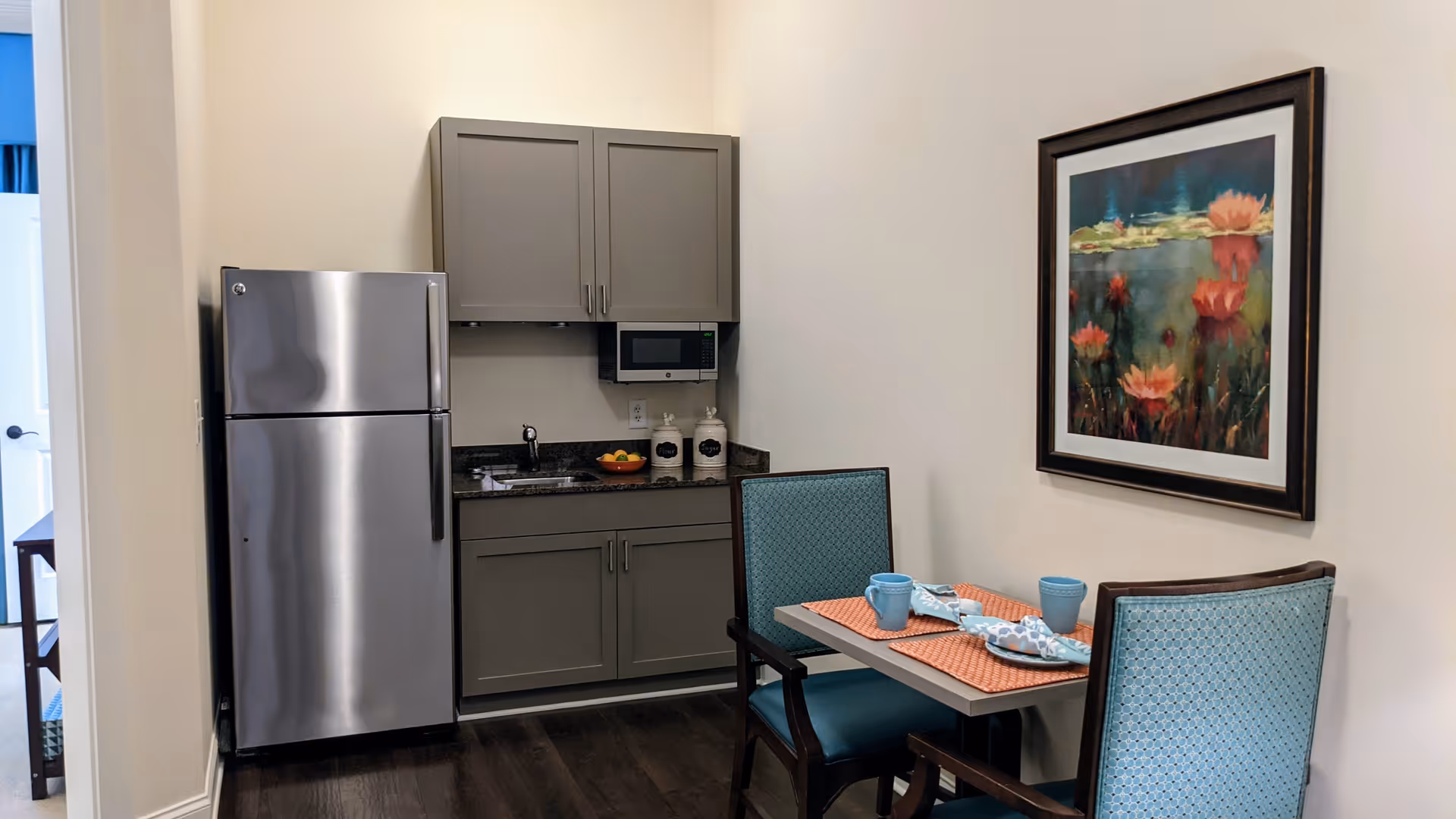 A small kitchen area with gray cabinets, a stainless steel refrigerator, a microwave, and a granite countertop with a sink. Next to the kitchen is a dining table set for two with blue chairs, orange placemats, blue cups, and napkins. A framed painting of water lilies hangs on the wall above the dining table.
