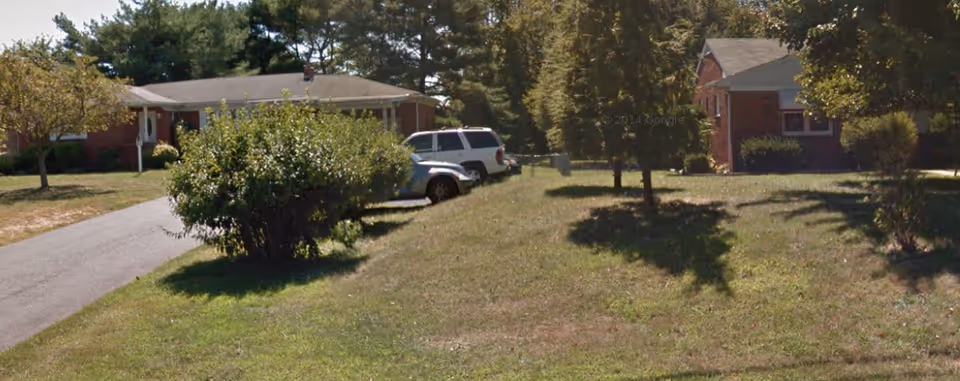 View of a senior care facility with a grassy front yard, several trees, a driveway with parked cars, and single-story brick buildings in the background under a clear sky.