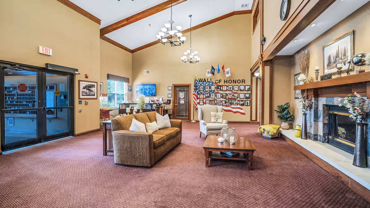 Spacious senior living lobby with sofas, armchairs and coffee table facing a fireplace and a 'Wall of Honor' photo display near the entrance.
