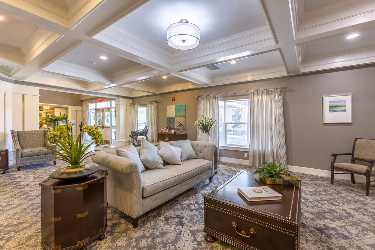 A well-lit living room area in a senior living facility featuring a beige tufted sofa with multiple cushions, a dark wooden coffee table with decorative items, a side table with a green plant, and additional seating including an armchair and a wooden chair. The room has a coffered ceiling with recessed lighting, large windows with curtains, and neutral-colored walls with framed artwork.