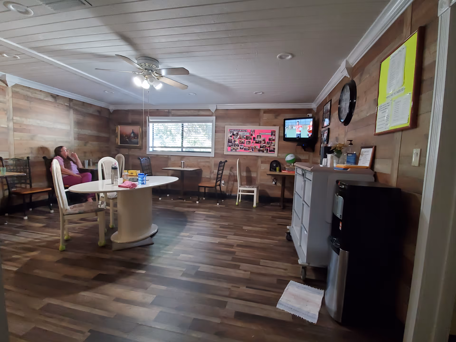 Communal senior living room with a central table, chairs, a wall-mounted TV and a person seated at left.