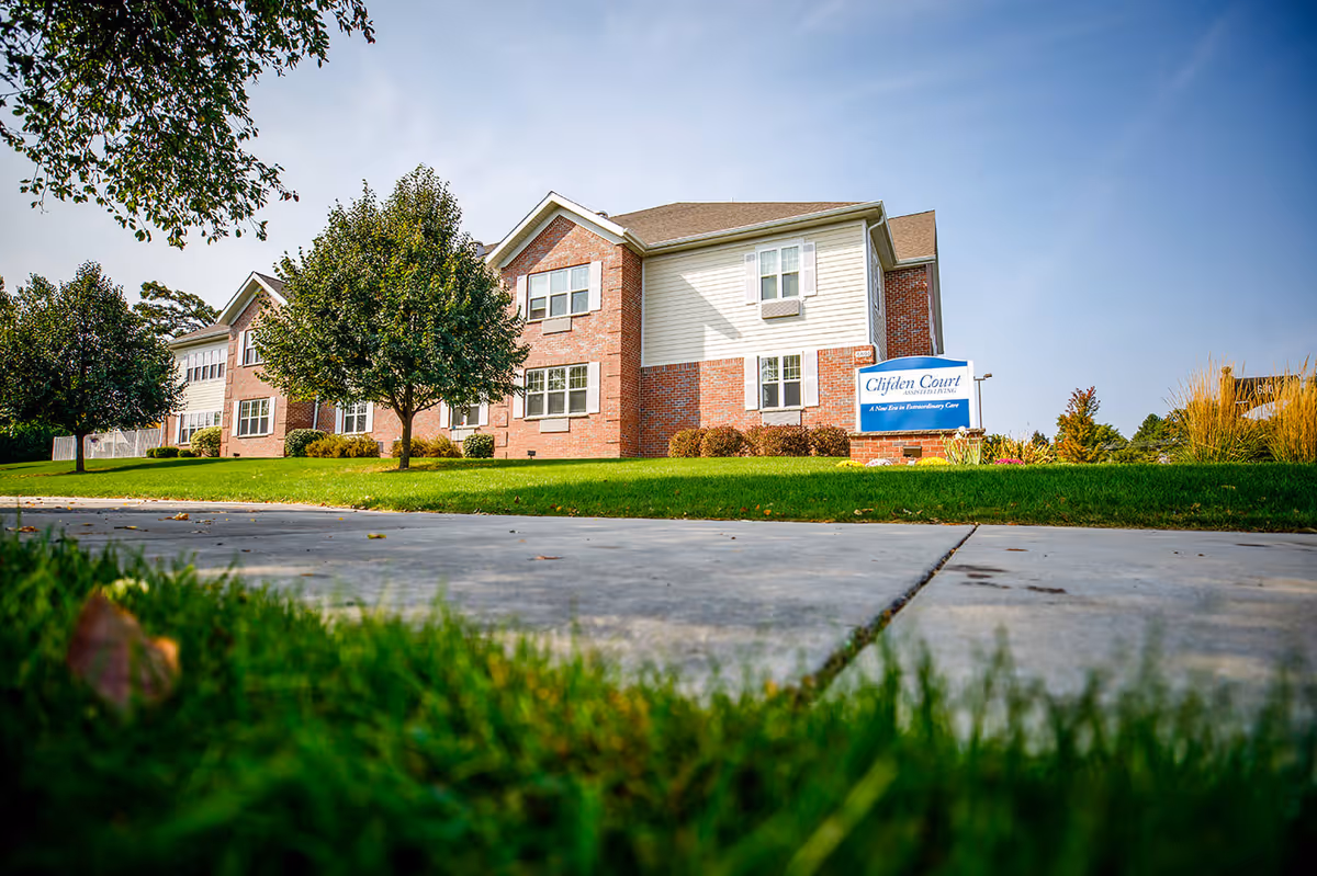 Exterior view of a two-story assisted living facility building with brick and siding facade, surrounded by green grass and trees under a clear blue sky. A sign in front reads 'Clifden Court Assisted Living, A New Era in Extraordinary Care.'