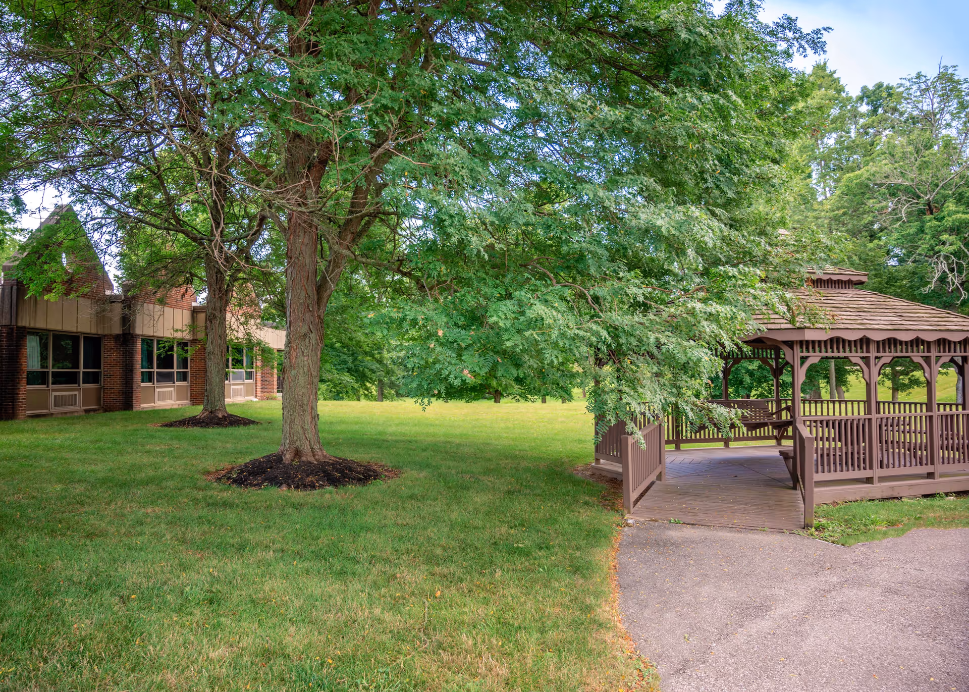 A green outdoor area with a large tree in the center, a brown wooden gazebo on the right, and a brick building with windows on the left. The scene is lush with grass and surrounded by more trees in the background.