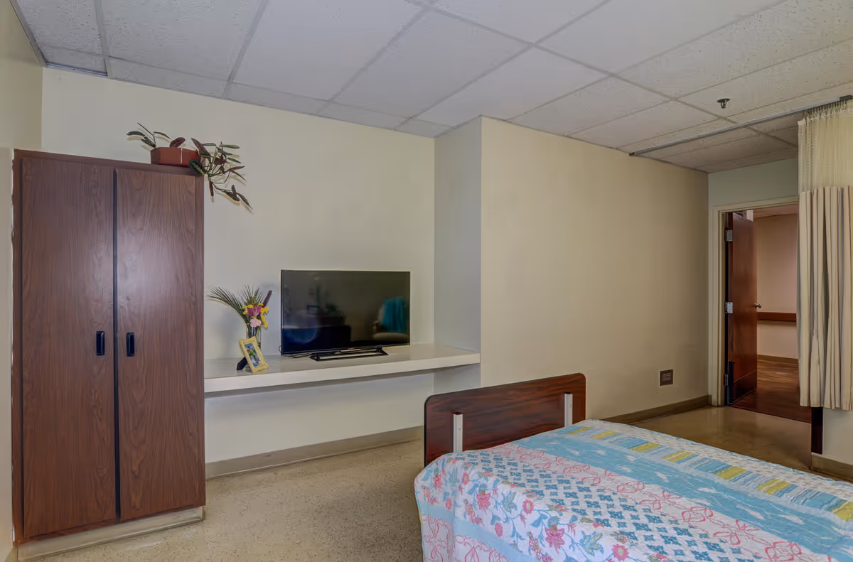 A simple bedroom in a senior living facility with a single bed covered by a colorful quilt, a wooden wardrobe with a potted plant on top, a flat-screen TV on a white wall-mounted shelf, and an open door leading to a hallway. The room has a drop ceiling and beige walls.