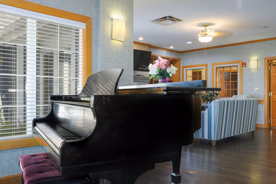Black grand piano in a bright lounge area with upholstered seating, a vase of flowers, and large window blinds.