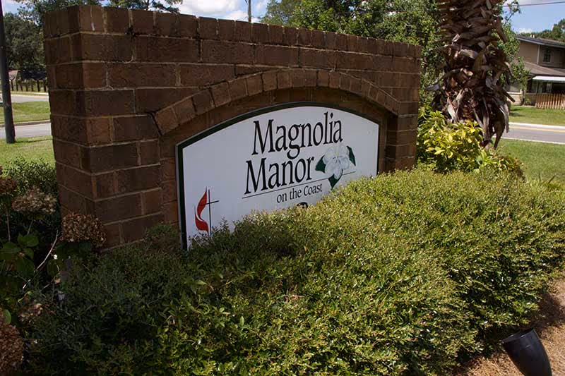 A brick sign surrounded by green bushes and plants displaying the name 'Magnolia Manor on the Coast' with a magnolia flower graphic and a red cross symbol.
