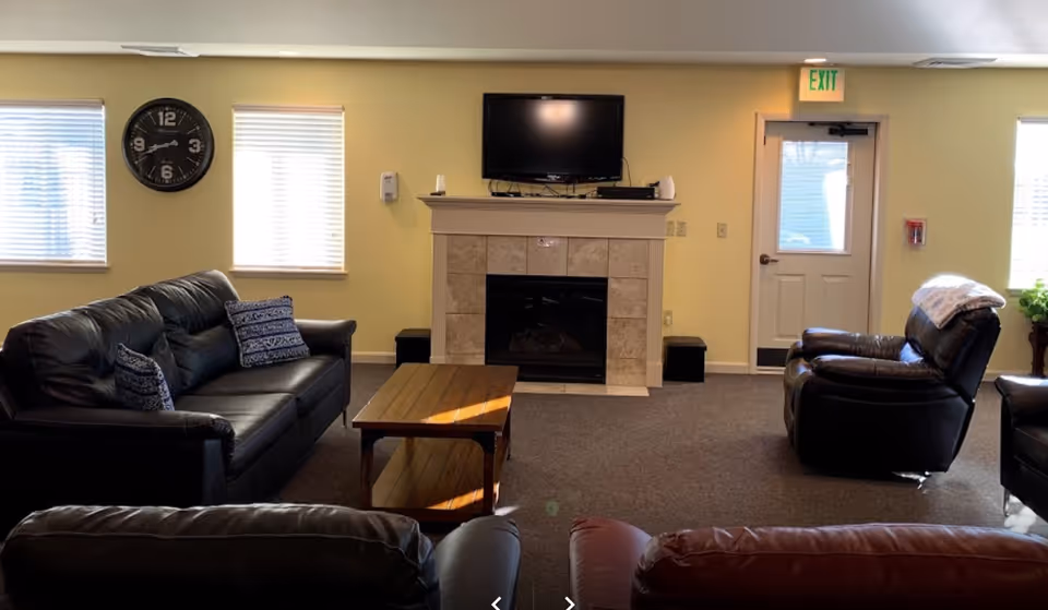 Bright common room with leather sofas and armchairs facing a fireplace topped by a wall-mounted TV, with windows and an exit door.