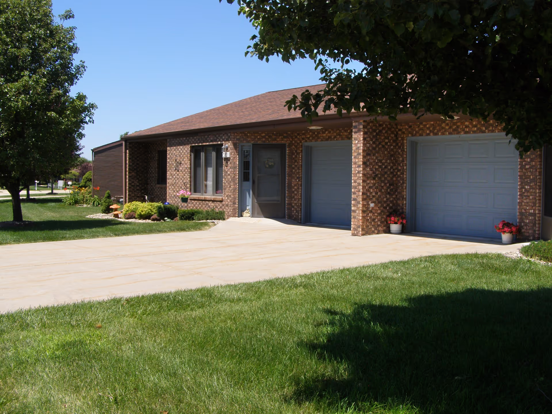 Exterior view of a single-story brick building with a brown roof, two closed garage doors, a front door with a small window, and a well-maintained lawn with trees and flower pots near the entrance.