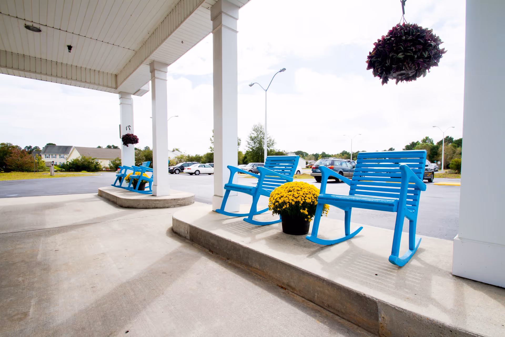 Covered outdoor seating area with bright blue rocking chairs and benches, yellow potted flowers, and hanging flower baskets, overlooking a parking lot with cars and greenery in the background.