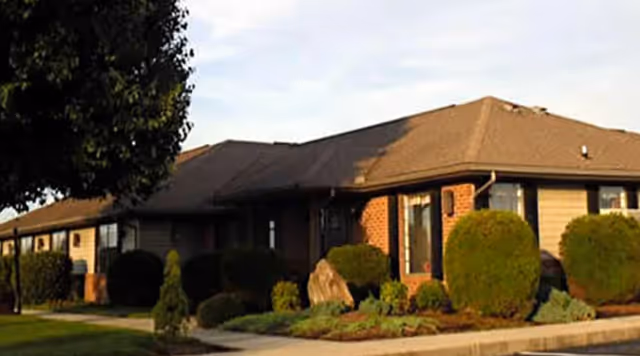 Front exterior of a single-story brick and siding assisted living building with trimmed shrubs, a tree, and a sidewalk.