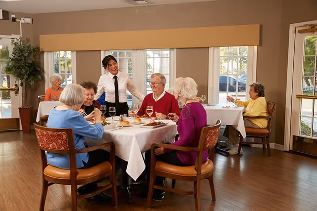 A group of elderly people sitting around a dining table in a well-lit room with large windows. A smiling waitress stands next to the table serving food. The table is set with plates of food, glasses of water, and bread rolls. Other elderly individuals are seated at separate tables in the background.