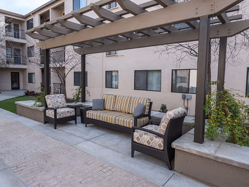 A shaded outdoor courtyard seating area with a pergola, striped sofa and two patterned armchairs in front of an apartment building.