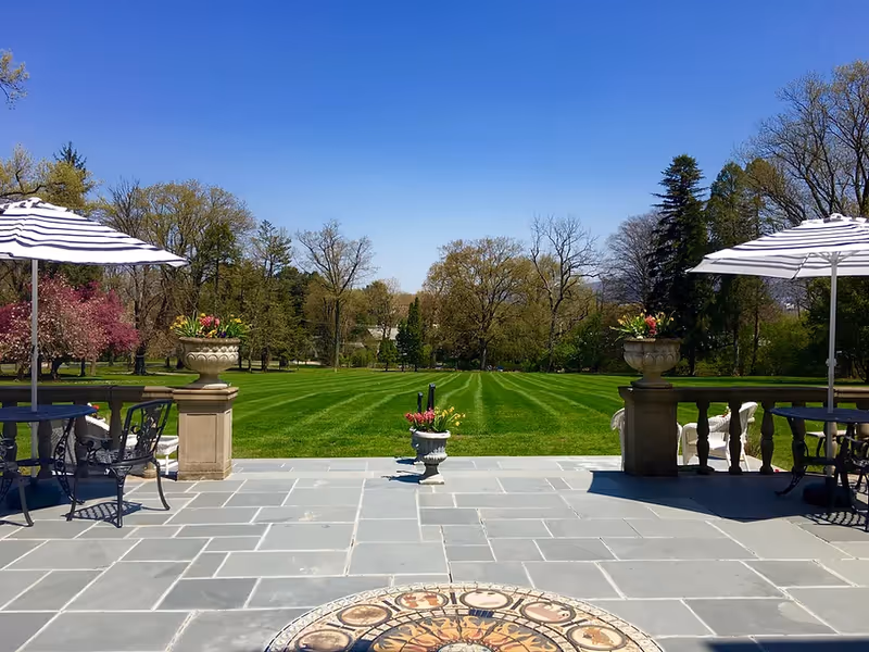 Stone patio with tables, striped umbrellas and planters overlooking a manicured lawn and trees under a clear blue sky.