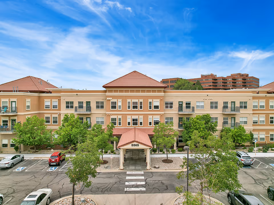 Front exterior view of a three-story senior living facility named The Inn at Greenwood Village with a covered entrance, multiple windows, balconies, trees, and a parking lot with several cars under a blue sky with some clouds.