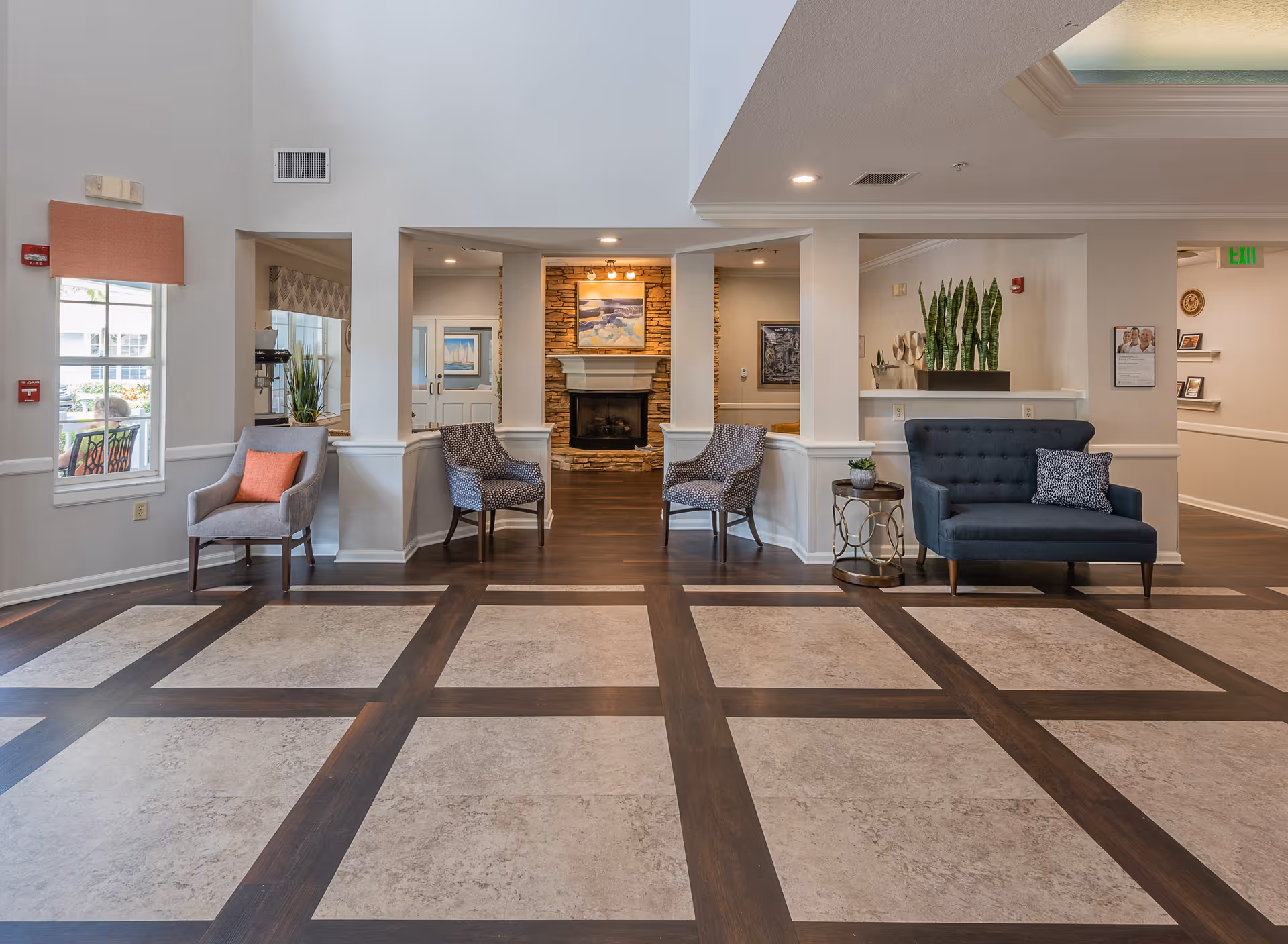 A spacious and well-lit common area in a senior living facility featuring a patterned floor with large square tiles bordered by dark wood. The room includes several chairs and a small sofa with cushions arranged along the walls. In the background, there is a stone fireplace with a painting above it, flanked by two armchairs. Potted plants and decorative items are placed on side tables and shelves, and windows allow natural light to enter the space.