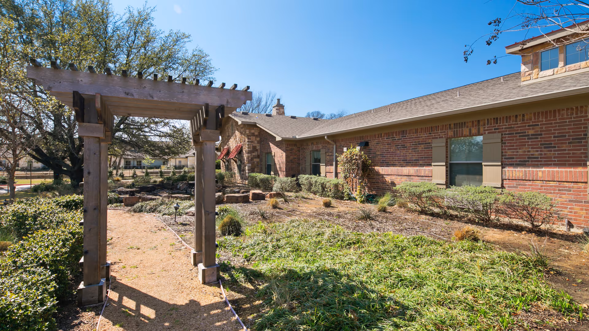 Outdoor garden area at Rosewood Assisted Living & Memory Care featuring a wooden pergola over a gravel pathway, surrounded by green shrubs, plants, and a brick building with windows and red awnings in the background under a clear blue sky.