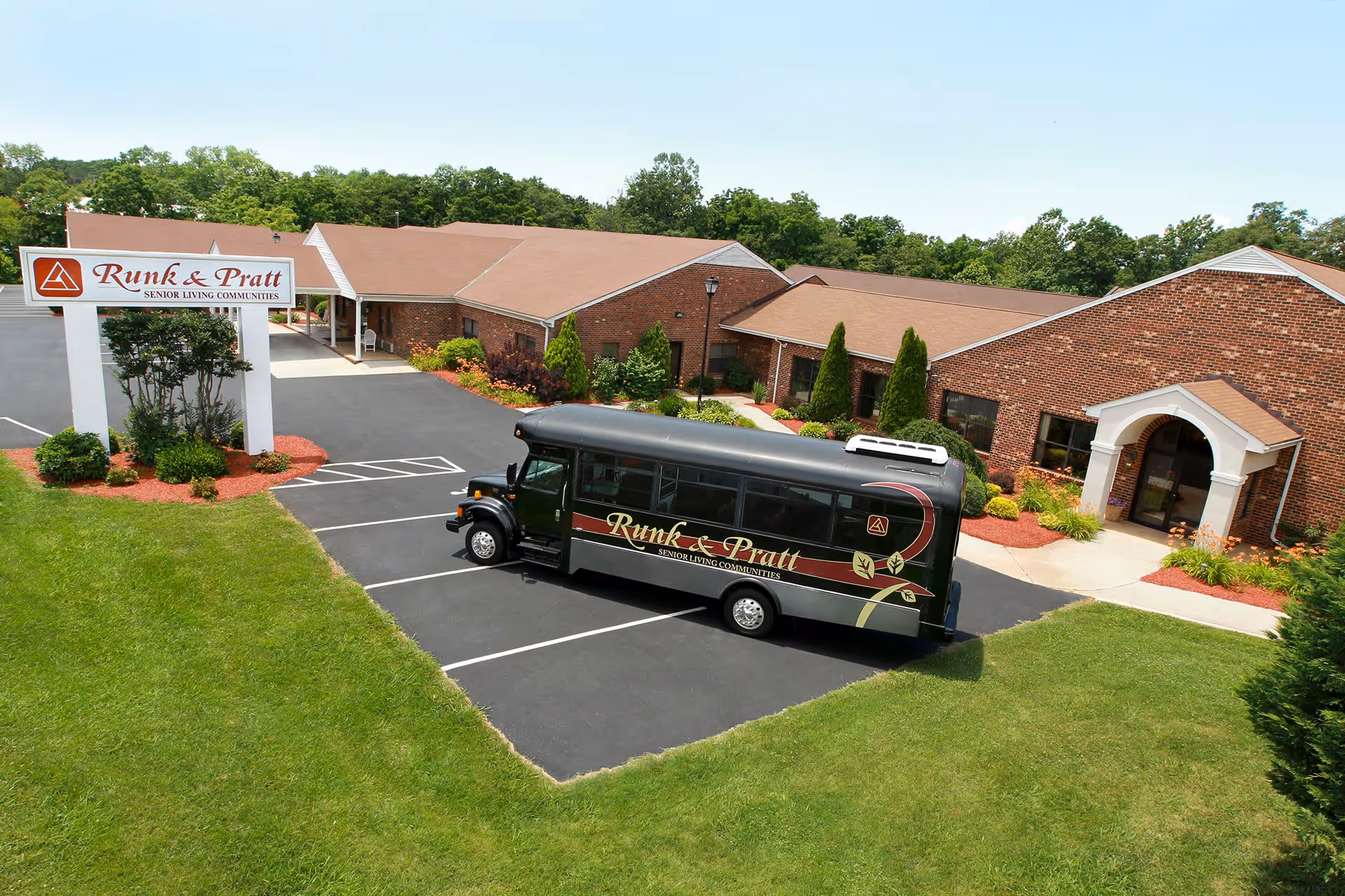 Exterior view of the Runk & Pratt senior living community building with a black shuttle bus parked in the lot. The building is made of brick with a tan roof, surrounded by green grass and landscaping. A large sign with the facility name is visible near the parking area.