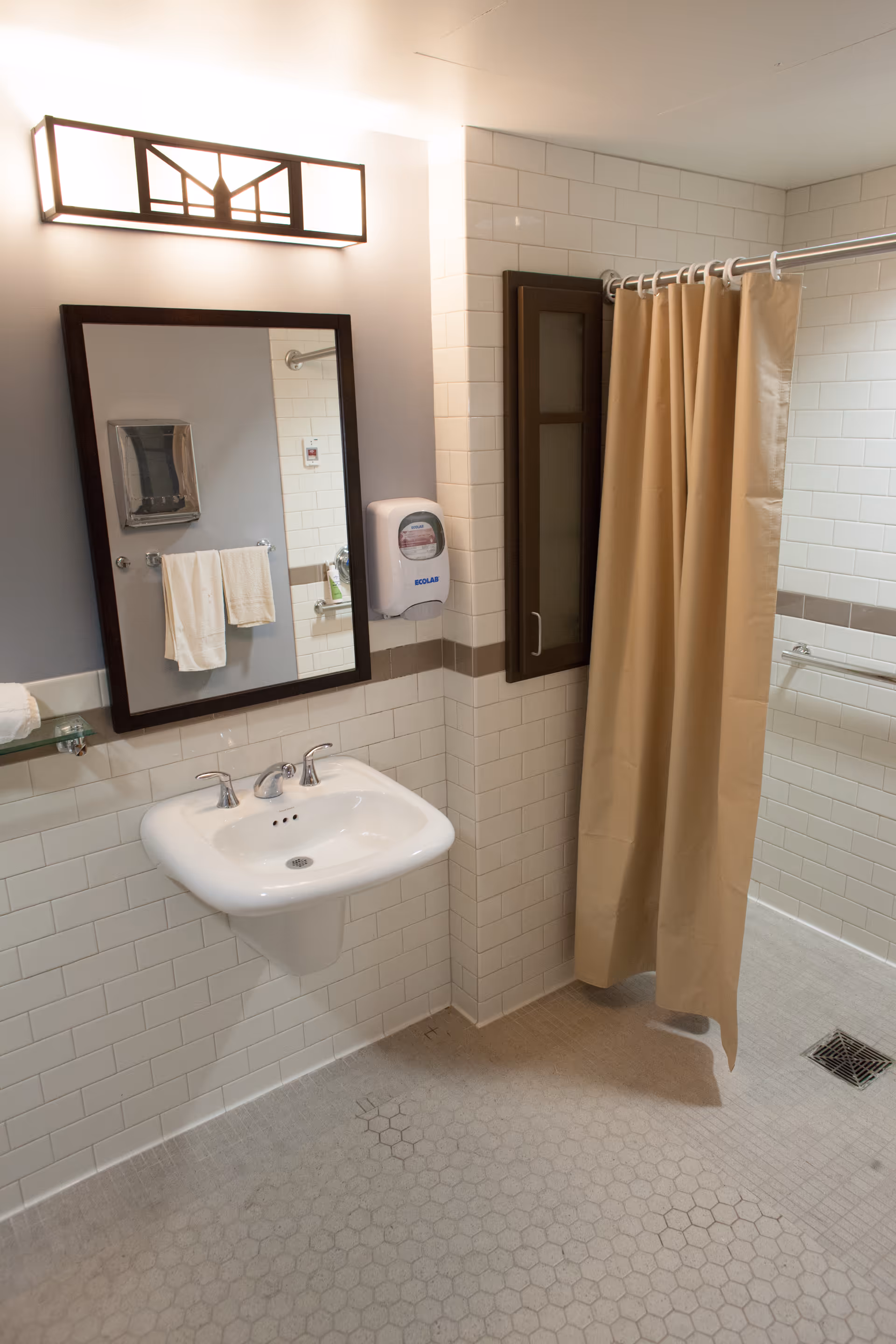 A clean bathroom with white tiled walls and hexagonal floor tiles. There is a white wall-mounted sink with silver faucets below a rectangular mirror framed in dark wood. Above the mirror is a decorative light fixture. To the right, there is a beige shower curtain hanging from a curved shower rod, partially covering a walk-in shower area. A soap dispenser is mounted on the wall next to the mirror, and a towel rack with white towels is visible in the mirror's reflection.