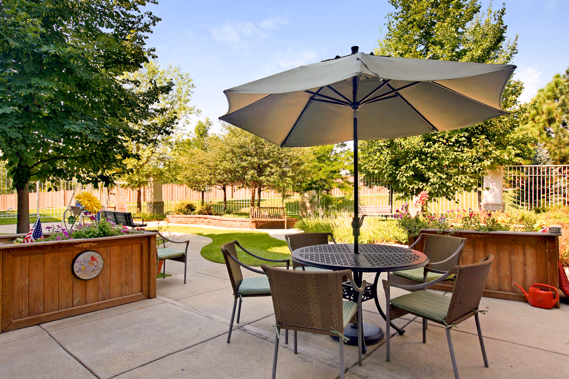 Outdoor patio area with a round metal table and four wicker chairs with cushions, shaded by a large umbrella. Surrounding the patio are wooden planter boxes with flowers, green trees, a curved concrete pathway, and benches in a garden setting under a clear blue sky.