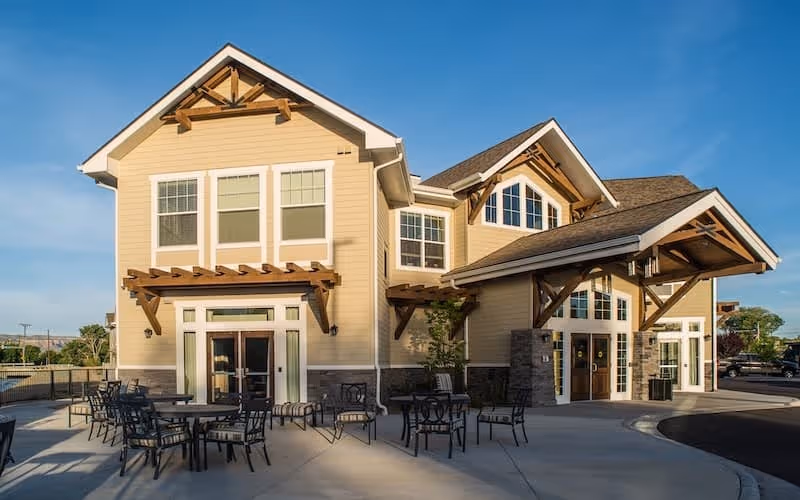 Exterior view of a two-story senior living facility building with beige siding, large windows, and wooden architectural details. There are several outdoor tables and chairs arranged on a concrete patio in front of the building under a clear blue sky.
