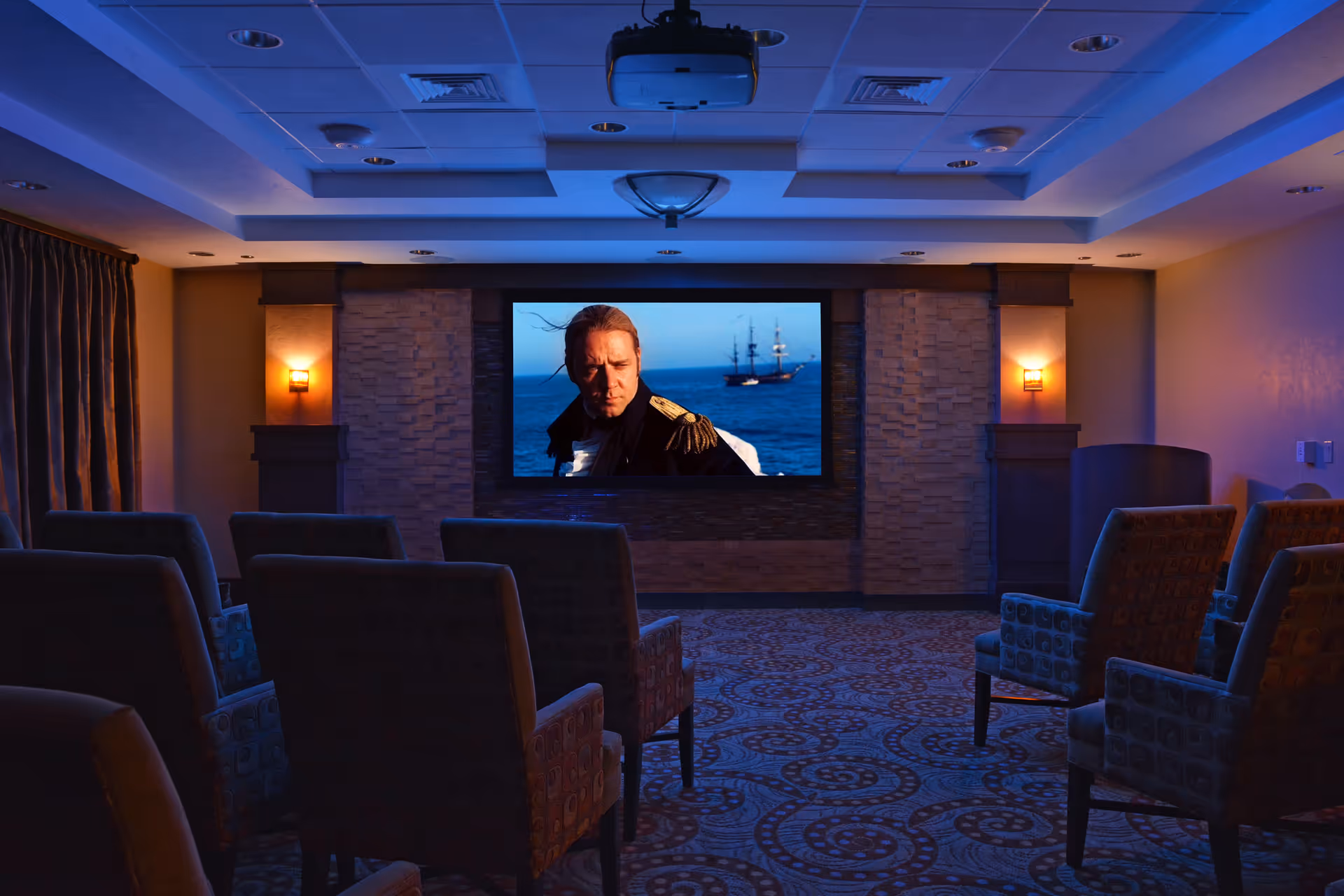 A small community theater/media room with upholstered chairs facing a large screen showing a seafaring movie scene.