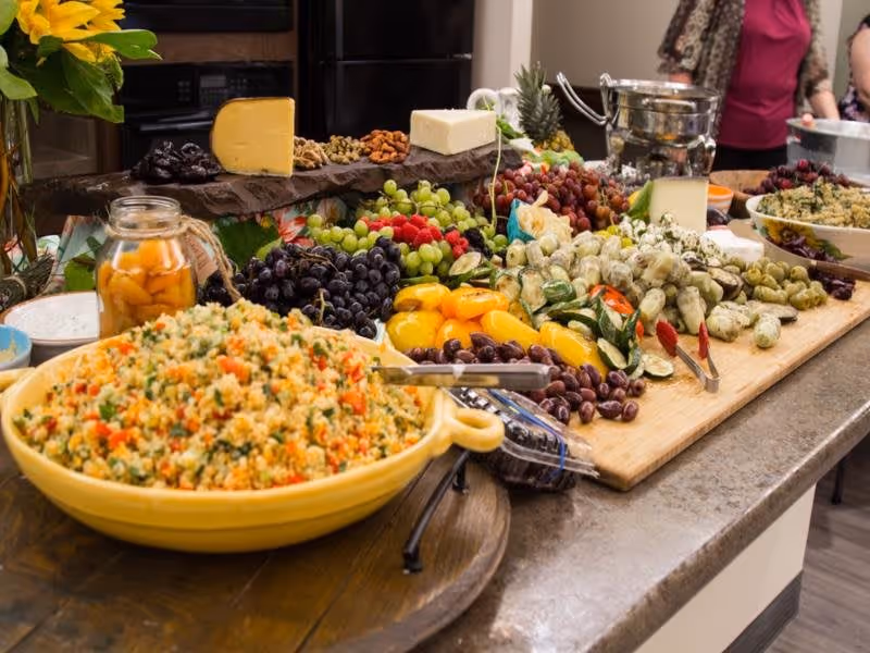 A buffet spread on a kitchen counter featuring a large bowl of mixed vegetable couscous, assorted cheeses, grapes, olives, pickles, nuts, and other fresh fruits and vegetables. Two people are partially visible in the background near the counter.