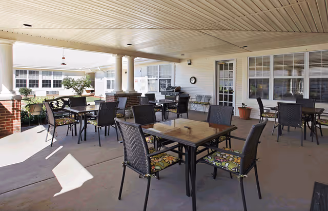 Covered outdoor patio area with several square tables and chairs with floral cushions, white pillars, a barbecue grill, and potted plants near a building with large windows and a door.