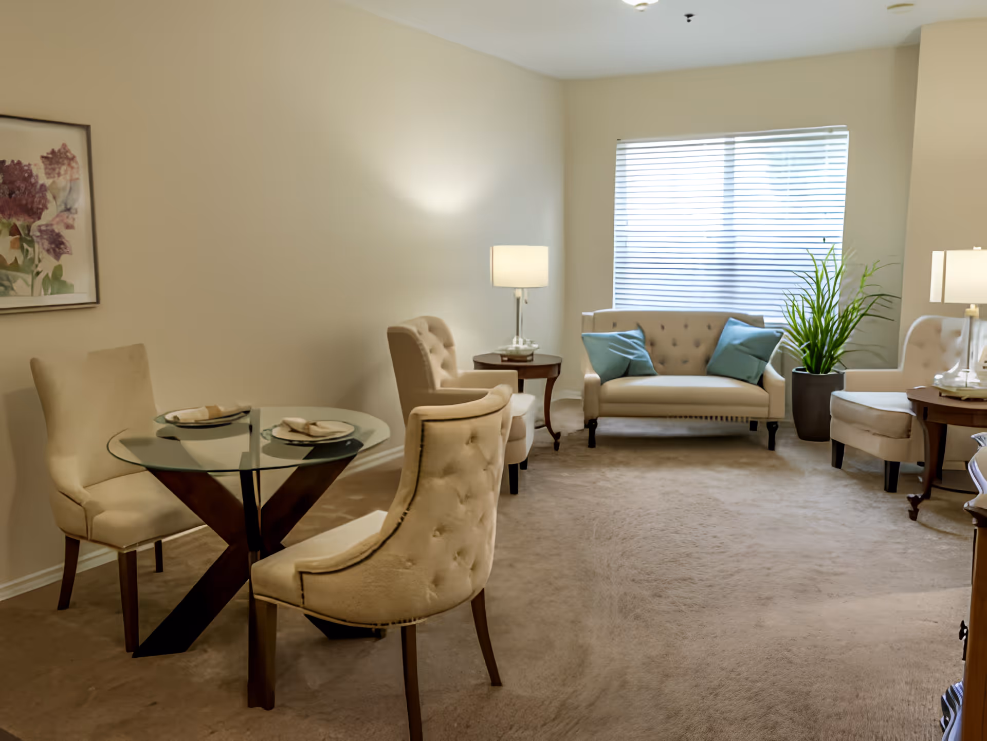 A cozy living room with beige carpet and walls, featuring a glass-top round table with two beige upholstered chairs, a beige loveseat with two blue cushions, two beige armchairs, two side tables with lamps, a large window with blinds, a potted plant, and a framed floral artwork on the wall.