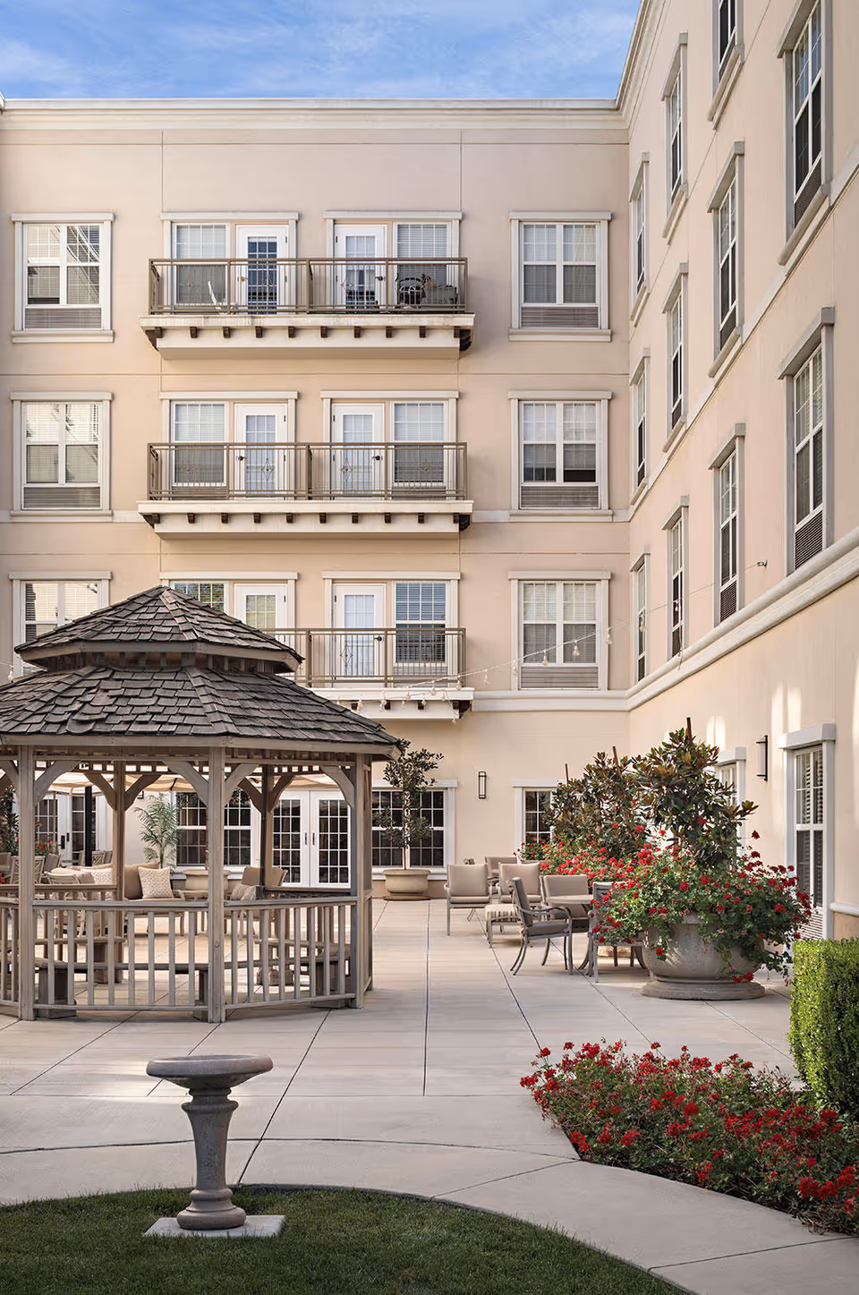 Outdoor courtyard area of a senior living facility featuring a wooden gazebo with a shingled roof, surrounded by patio seating with chairs and tables. The courtyard is landscaped with green grass, red flowering plants, and large potted plants. The beige multi-story building with balconies and windows encloses the courtyard under a clear blue sky.
