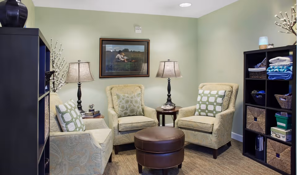 A cozy sitting area with three beige patterned armchairs arranged around a round brown leather ottoman. Each chair has a decorative pillow. There are two side tables with lamps and books, and a framed picture on the light green wall behind the chairs. On either side of the seating area are black shelving units holding baskets, folded blankets, and other items.