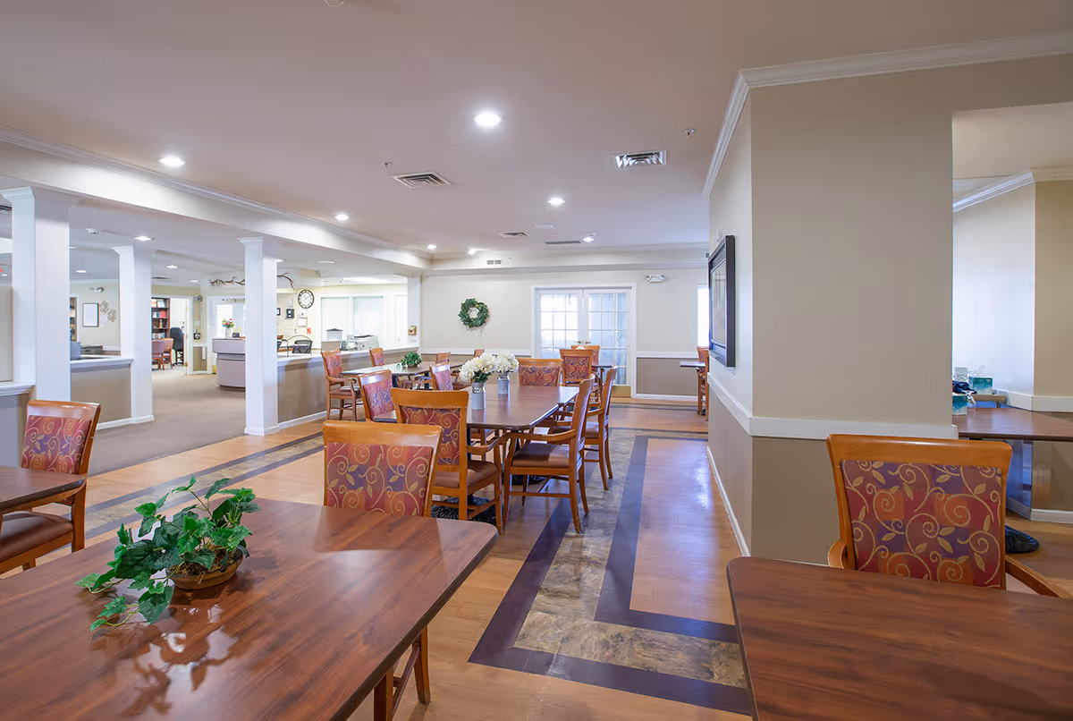 A bright and spacious dining area in a senior living facility with wooden tables and chairs featuring patterned upholstery. The room has light-colored walls, recessed ceiling lights, and decorative plants on the tables. In the background, there is a reception desk and a wall clock.