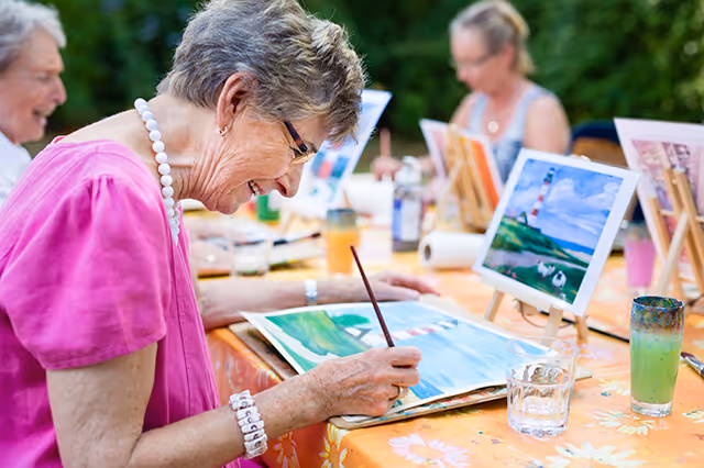Elderly women sitting at a table outdoors, engaged in painting on canvases with paintbrushes. One woman in the foreground is smiling and focused on her artwork, which depicts a landscape scene with water and greenery. The table is covered with a floral tablecloth and has glasses of water and paint supplies.