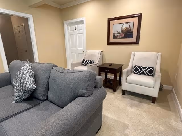 A cozy living room with a gray sofa featuring patterned cushions, two beige armchairs with black and white patterned pillows, a small dark wooden side table between the armchairs, beige carpet, and a framed picture of a basket on the wall.