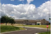 Wide single-story beige retirement community building under a partly cloudy sky with a road, grassy lawn and a stop sign in the foreground.