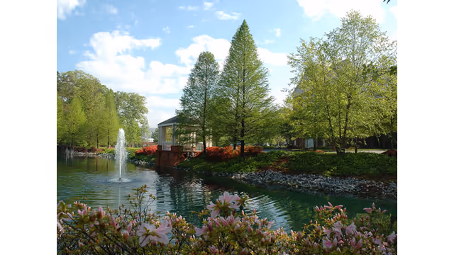A landscaped pond with a central fountain, blooming flowers and trees, and a small pavilion under a blue sky.
