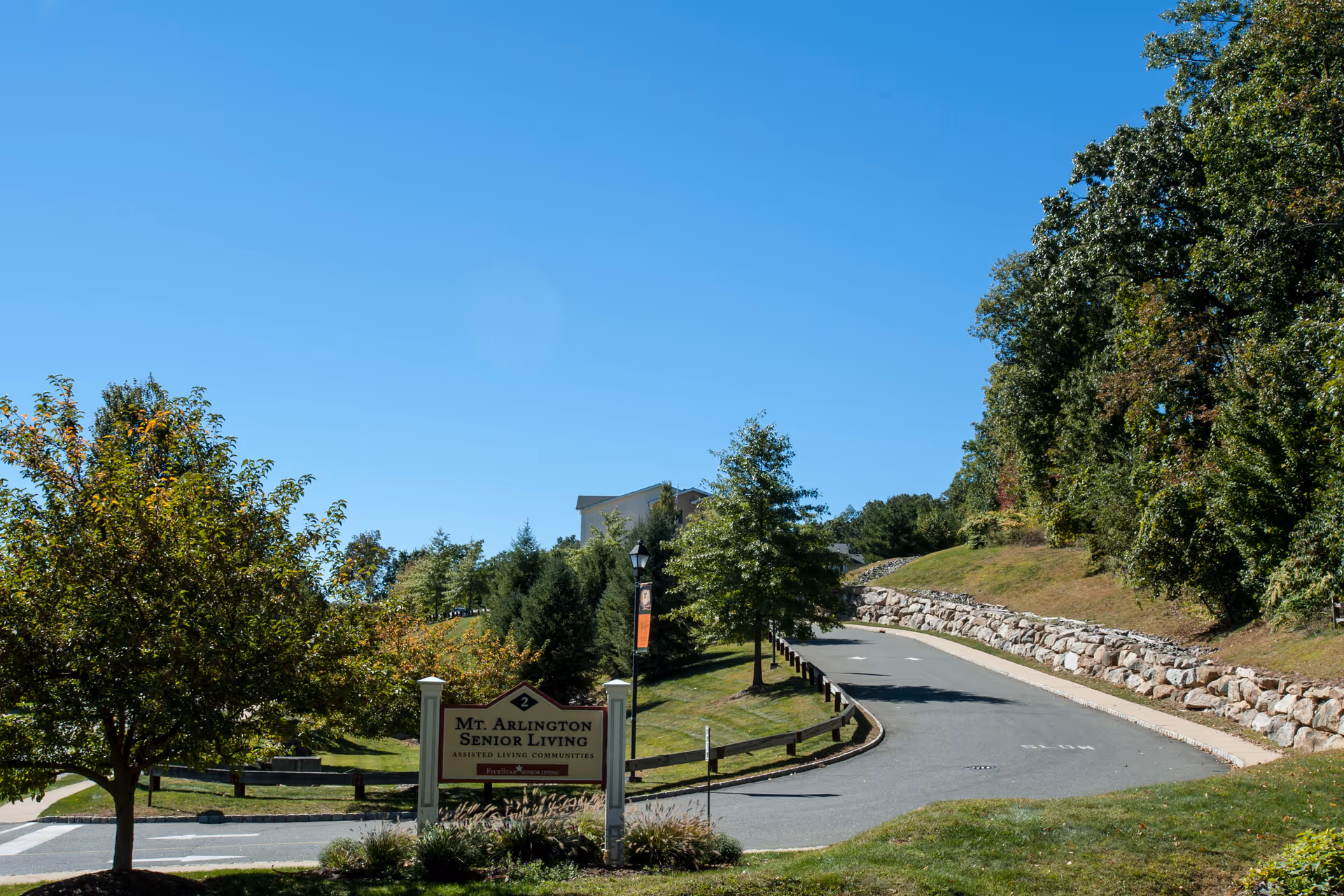 A paved driveway leading uphill surrounded by green trees and grass under a clear blue sky. A sign at the entrance reads 'Mt. Arlington Senior Living Assisted Living Communities.'