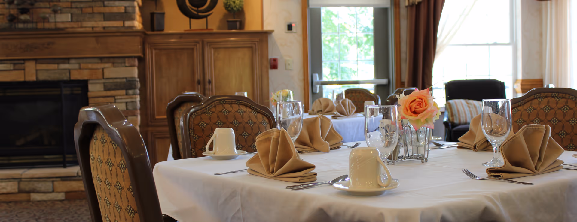 A dining room table set with beige folded napkins, white coffee cups on saucers, clear water glasses, and silverware. A small vase with a pink rose is placed in the center of the table. The background shows upholstered chairs, a stone fireplace, wooden cabinetry, and a door with glass panes letting in natural light.
